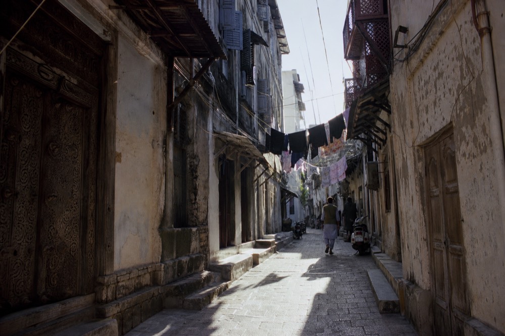 Narrow coral stone alley with carved wooden doors and shuttered balconies, laundry strung overhead, hard shadow and bright sun on the worn paving below