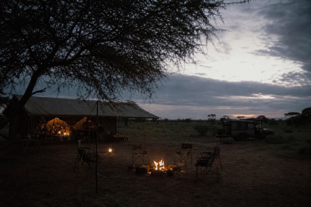 Canvas tented camp at dusk with lantern light spilling from the mess tent, folding chairs around a low fire ring, dark plains behind