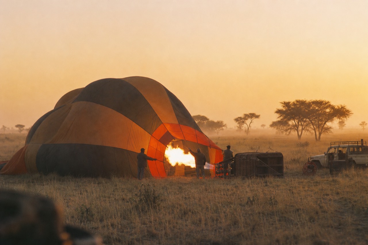 A half inflated balloon envelope lying on pale dawn grass, ground crew silhouettes holding the mouth open, burner flame lighting the fabric orange from inside
