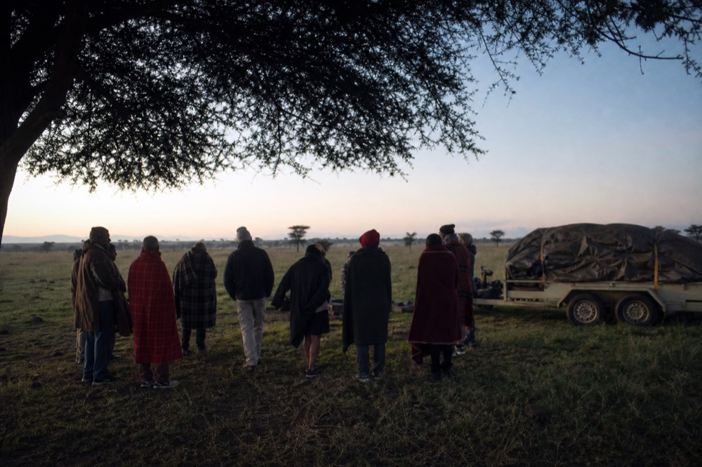 Passengers standing in a loose group on open grass in cold half-light before sunrise, blankets over shoulders, a folded balloon envelope on a trailer behind them