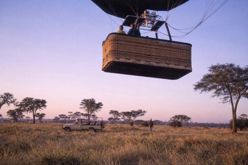 Underside of a balloon basket lifting away from the ground, woven wicker and burner frame silhouetted against a pale lilac dawn sky