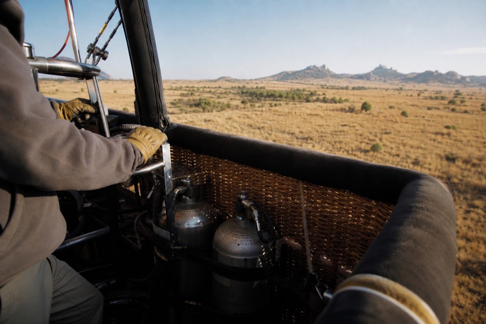 A pilot's gloved hands on the burner control frame inside a balloon basket, steel fuel cylinders strapped upright against the padded wicker wall