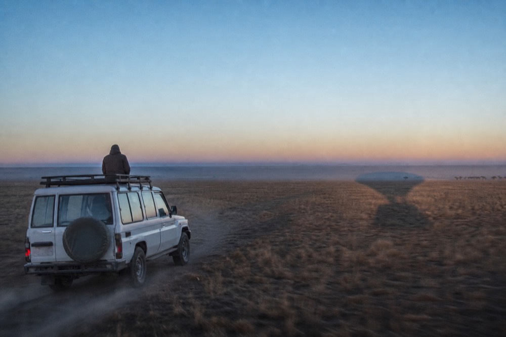 A white four-wheel-drive chase vehicle trailing dust across open plains, following the dark oval shadow of a balloon on the grass ahead