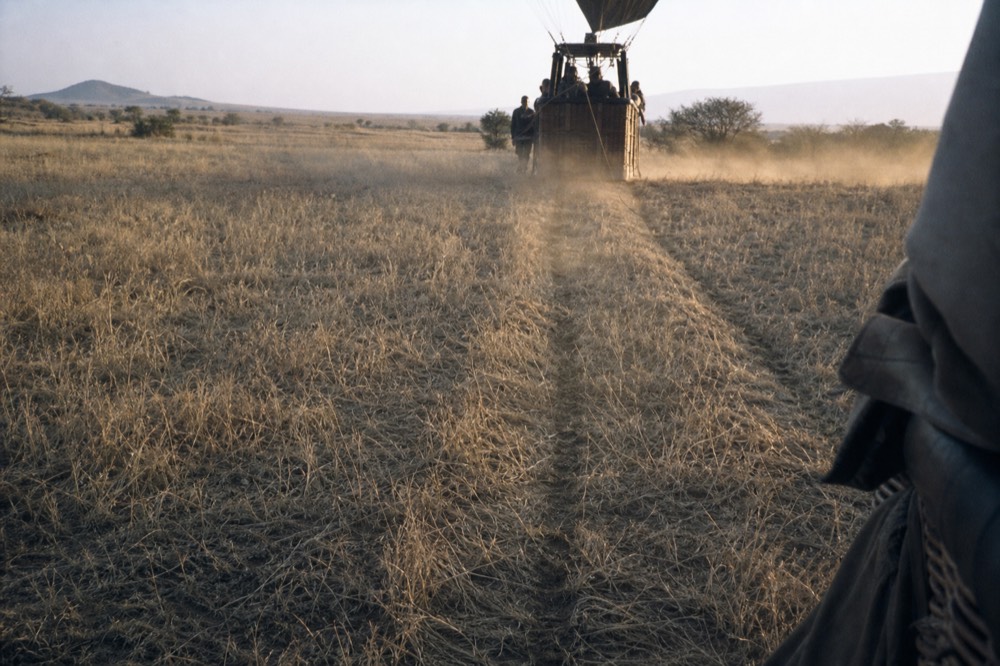 Two parallel drag marks pressed through pale dry grass where a basket touched down, dust still hanging in low sunlight, folded fabric edge visible at the frame edge