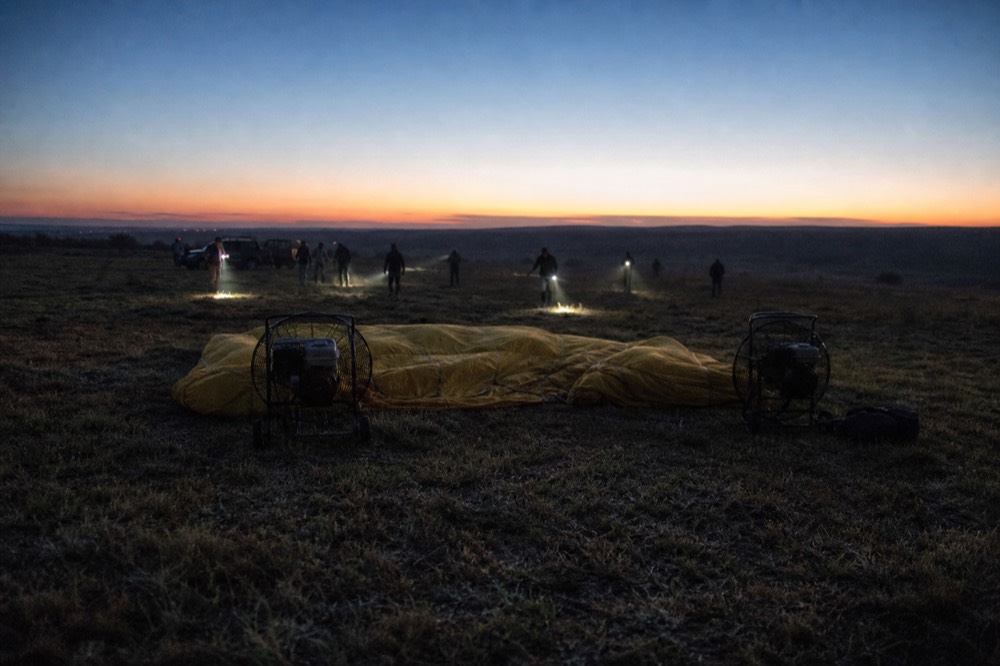 Two petrol fans pushing cold air into a slack unbranded envelope on dark grass before dawn, crew silhouettes moving through crossed headlamp beams
