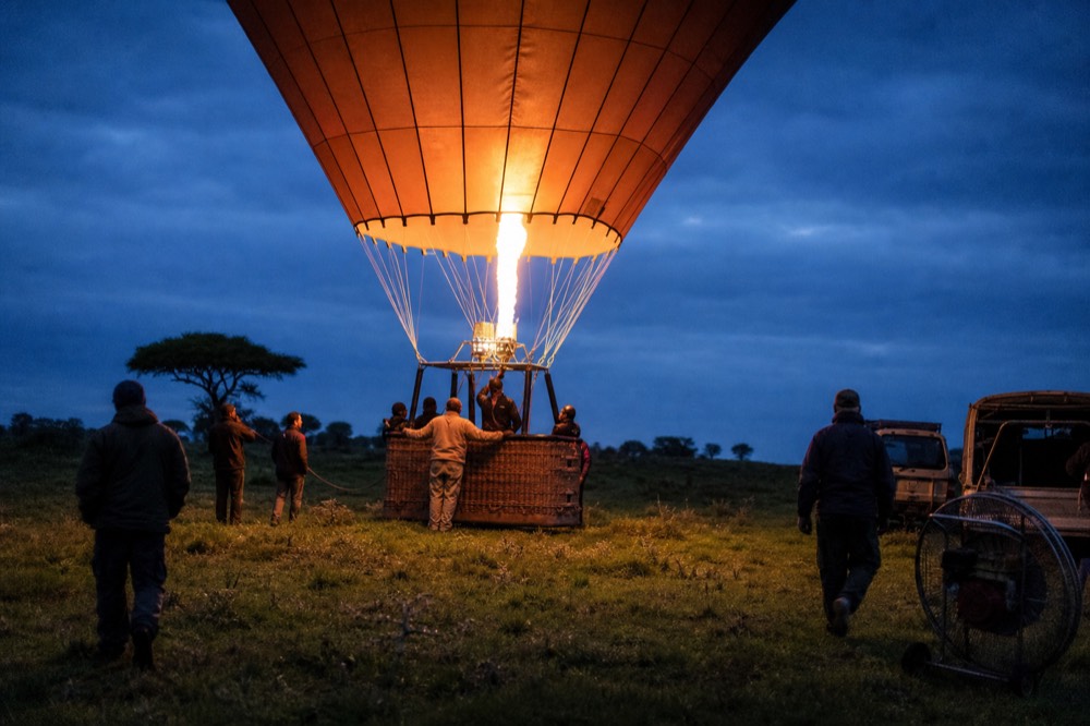 Burner flame glowing inside the crown of a plain orange envelope against a deep blue pre-sunrise sky, ropes taut and crew hands on the basket edge