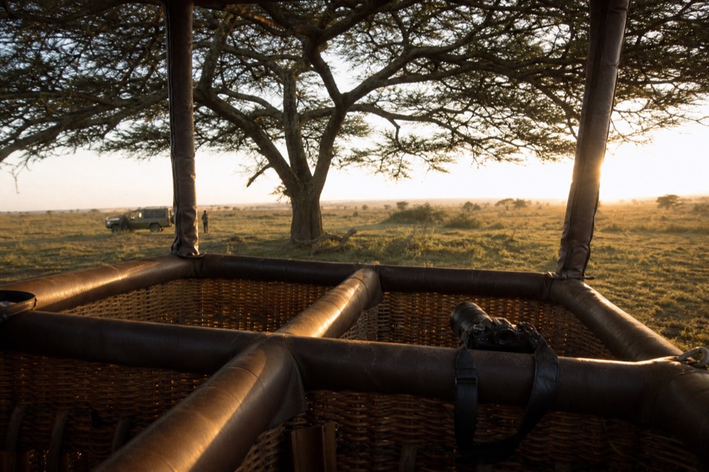 Interior of a woven wicker balloon basket showing padded rails and separate standing compartments, a camera strap looped over a rail, warm sunrise light across the weave